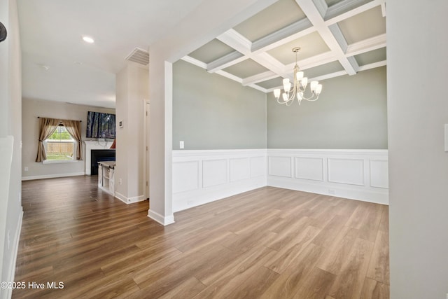 empty room with a chandelier, wood finished floors, beamed ceiling, coffered ceiling, and a wainscoted wall