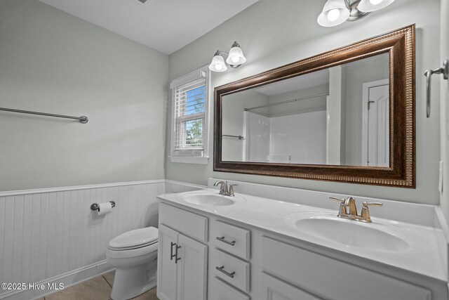 full bathroom with toilet, a wainscoted wall, double vanity, and a shower