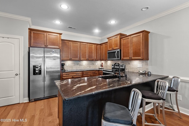 kitchen with appliances with stainless steel finishes, a peninsula, brown cabinetry, dark stone countertops, and light wood finished floors