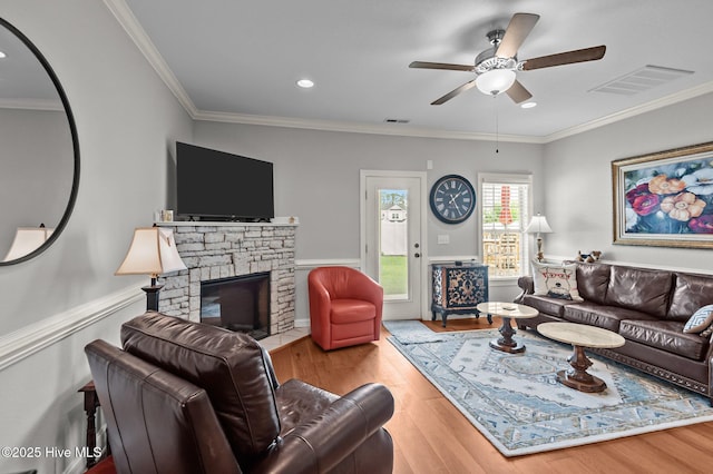 living room featuring wood finished floors, ceiling fan, a stone fireplace, ornamental molding, and wainscoting