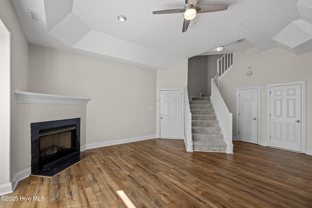 unfurnished living room featuring stairs, wood finished floors, a ceiling fan, baseboards, and a fireplace with raised hearth