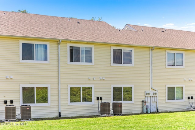 rear view of property with cooling unit, a lawn, and roof with shingles