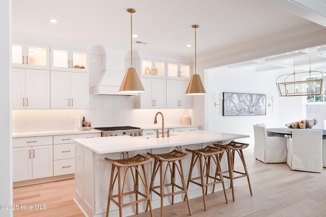 kitchen with light wood finished floors, premium range hood, a kitchen island with sink, light countertops, and white cabinetry