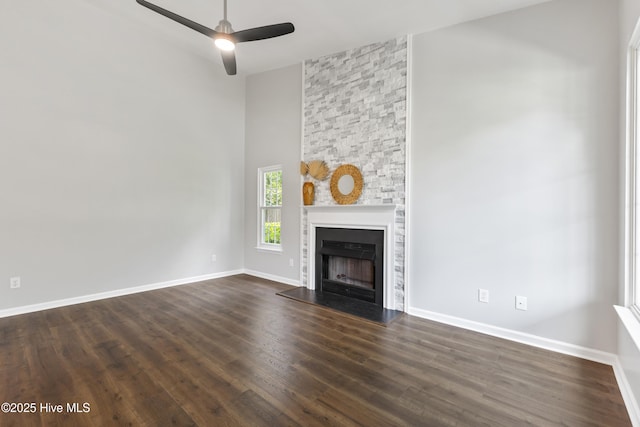 unfurnished living room featuring baseboards, a ceiling fan, dark wood-type flooring, a stone fireplace, and a high ceiling