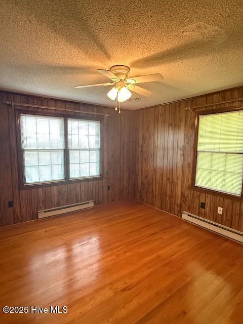 empty room featuring a baseboard heating unit, a textured ceiling, wood finished floors, wooden walls, and a ceiling fan