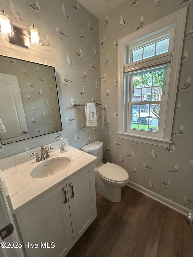 bathroom featuring wood finished floors, wallpapered walls, toilet, vanity, and baseboards