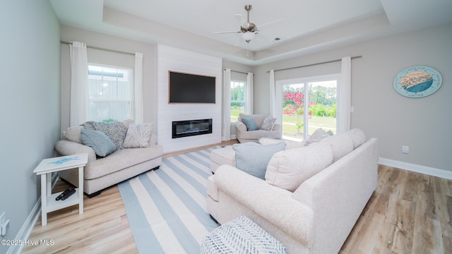 living area featuring a tray ceiling, light wood-style flooring, baseboards, a ceiling fan, and a large fireplace