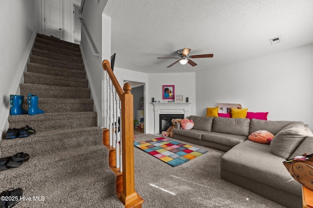 living room featuring a ceiling fan, stairway, a textured ceiling, a glass covered fireplace, and carpet floors
