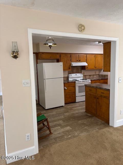 908 E 7th St, Lumberton, NC 28358 - kitchen with white appliances, under cabinet range hood, brown cabinetry, dark countertops, and dark colored carpet