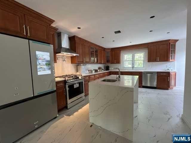 kitchen featuring sink, wall chimney exhaust hood, light stone countertops, an island with sink, and stainless steel appliances