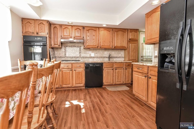 kitchen featuring backsplash, black appliances, light hardwood / wood-style flooring, a tray ceiling, and light stone counters