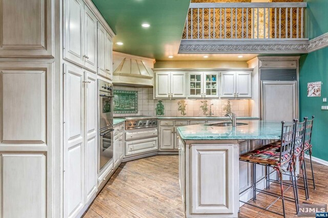 kitchen with light stone countertops, light wood-type flooring, custom range hood, sink, and an island with sink