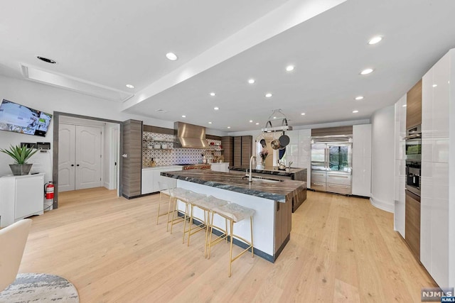 kitchen featuring light wood-type flooring, wall chimney exhaust hood, a large island with sink, white cabinetry, and a breakfast bar area