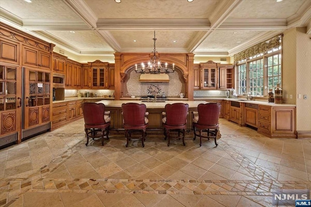 kitchen featuring coffered ceiling, backsplash, a large island with sink, pendant lighting, and a breakfast bar