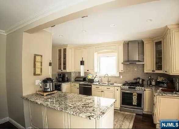 kitchen with light stone countertops, cream cabinets, stainless steel gas range oven, and wall chimney exhaust hood