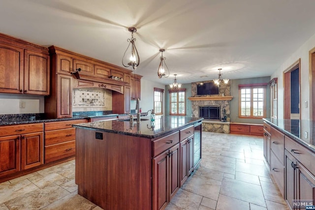 kitchen featuring dark stone counters, a stone fireplace, sink, hanging light fixtures, and an island with sink