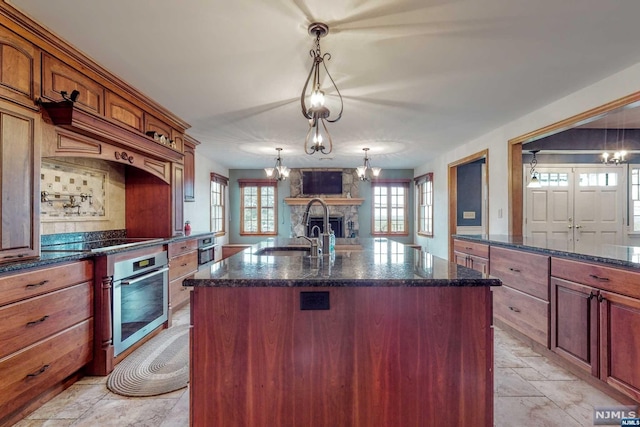 kitchen with stainless steel oven, sink, a stone fireplace, an island with sink, and pendant lighting