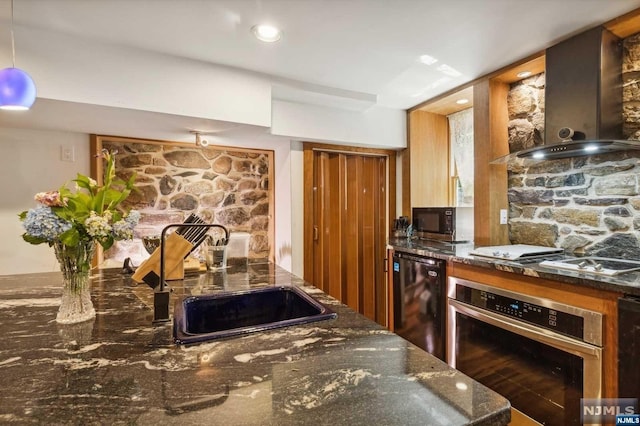 kitchen featuring sink, stainless steel appliances, wall chimney range hood, dark stone counters, and decorative light fixtures