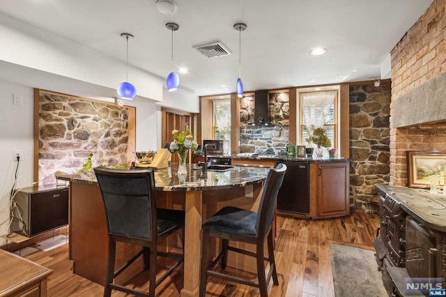 bar featuring dishwasher, wall chimney range hood, light hardwood / wood-style flooring, dark stone countertops, and decorative light fixtures