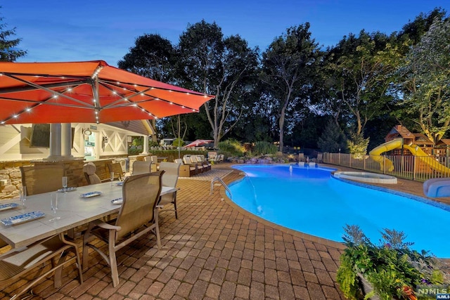 pool at dusk with a playground, a patio, and an outdoor bar