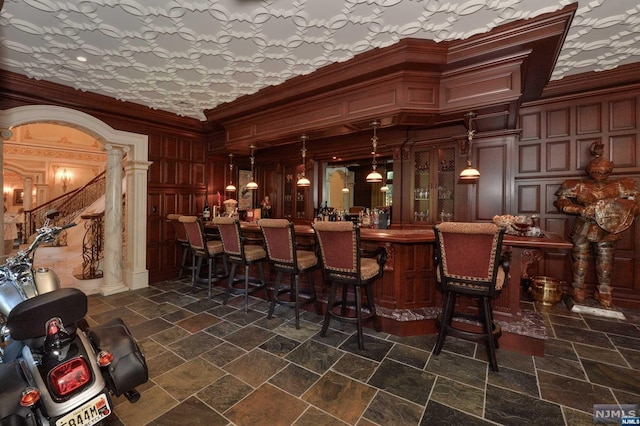 bar featuring pendant lighting, ornate columns, ornamental molding, and wood walls