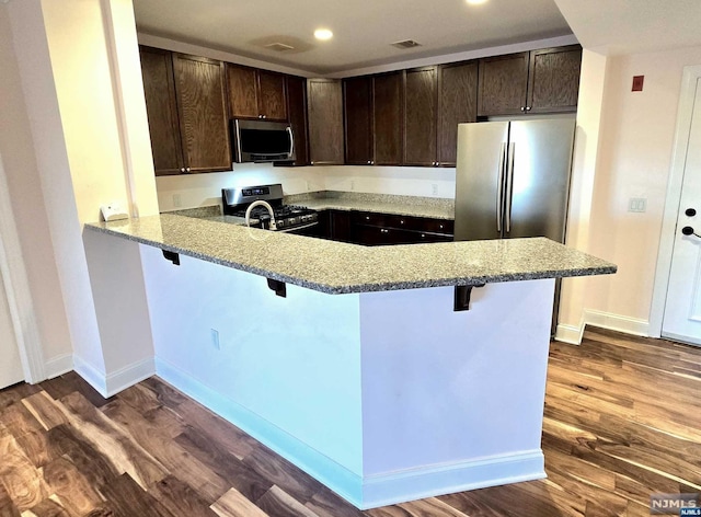 kitchen with a kitchen bar, kitchen peninsula, dark brown cabinetry, stainless steel appliances, and dark wood-type flooring