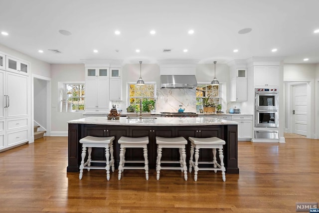 kitchen with double oven, dark wood-type flooring, a spacious island, wall chimney range hood, and pendant lighting