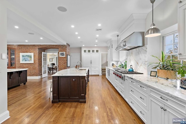 kitchen featuring wall chimney exhaust hood, an island with sink, pendant lighting, stainless steel gas stovetop, and light wood-type flooring