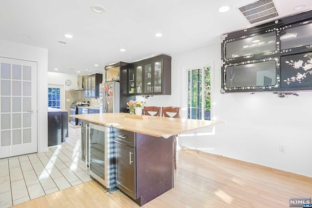 kitchen with dark brown cabinetry, wall chimney exhaust hood, light hardwood / wood-style flooring, a breakfast bar area, and appliances with stainless steel finishes