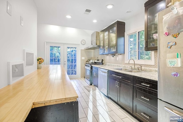 kitchen with light stone countertops, appliances with stainless steel finishes, french doors, sink, and wall chimney range hood