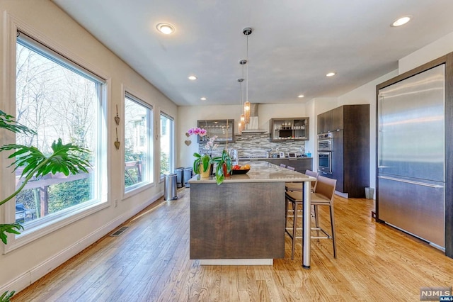 kitchen featuring decorative backsplash, light wood-type flooring, dark brown cabinets, wall chimney range hood, and pendant lighting