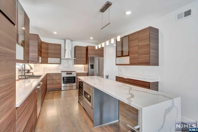 kitchen featuring stainless steel appliances, sink, wall chimney range hood, a kitchen island, and hanging light fixtures