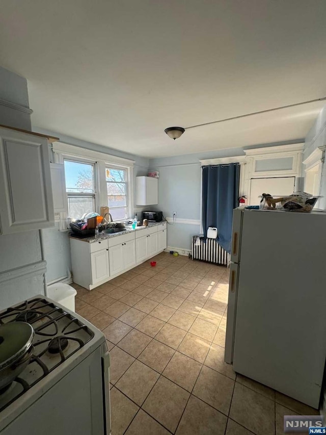 kitchen with radiator heating unit, sink, white cabinets, light tile patterned flooring, and white appliances