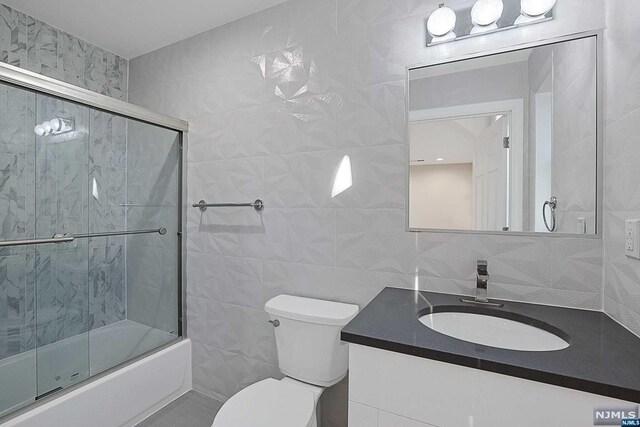 full bathroom featuring combined bath / shower with glass door, vanity, toilet, and tile walls