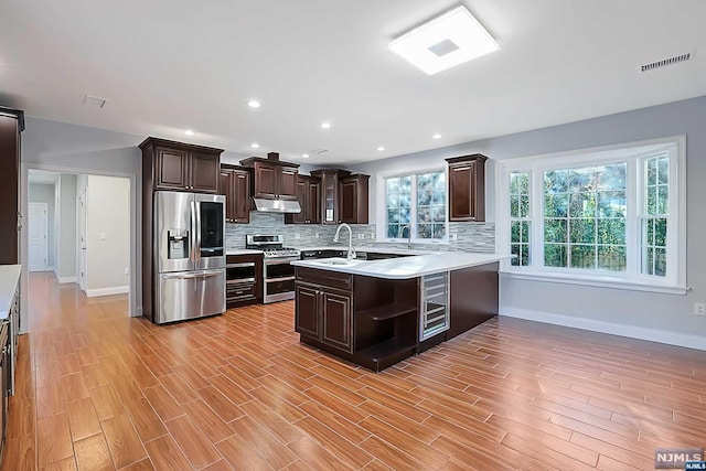 kitchen with kitchen peninsula, backsplash, dark brown cabinetry, stainless steel appliances, and light hardwood / wood-style floors