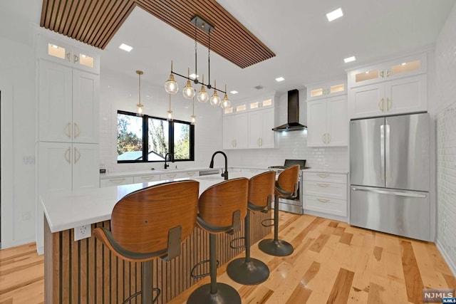 kitchen with wall chimney exhaust hood, stainless steel appliances, a center island with sink, light hardwood / wood-style flooring, and white cabinets