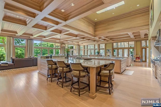 kitchen with light brown cabinets, a large island with sink, coffered ceiling, light hardwood / wood-style flooring, and light stone counters