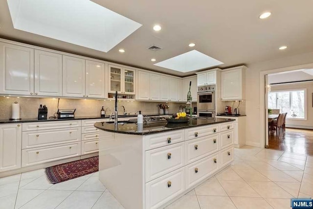 kitchen with a skylight, white cabinetry, a center island with sink, and light tile patterned floors