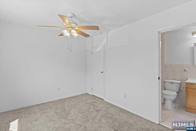 unfurnished bedroom featuring ensuite bathroom, tile walls, light colored carpet, and ceiling fan