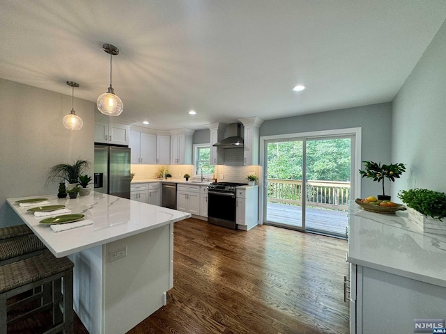 kitchen with dark wood-type flooring, wall chimney range hood, hanging light fixtures, white cabinetry, and stainless steel appliances