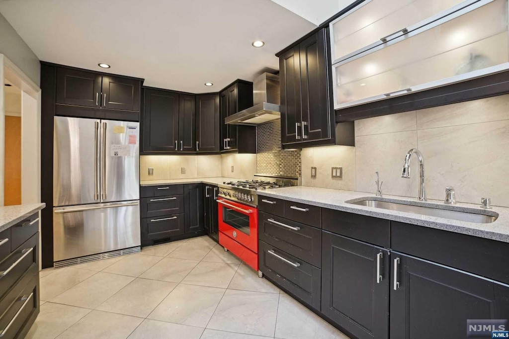 kitchen with high quality appliances, wall chimney range hood, sink, decorative backsplash, and light stone counters