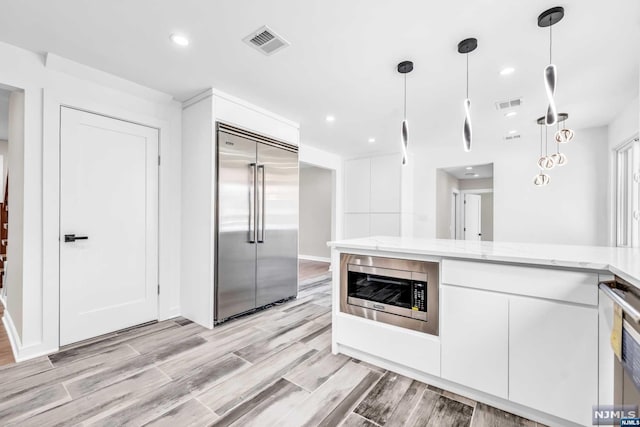 kitchen featuring pendant lighting, built in appliances, light stone countertops, light wood-type flooring, and white cabinetry