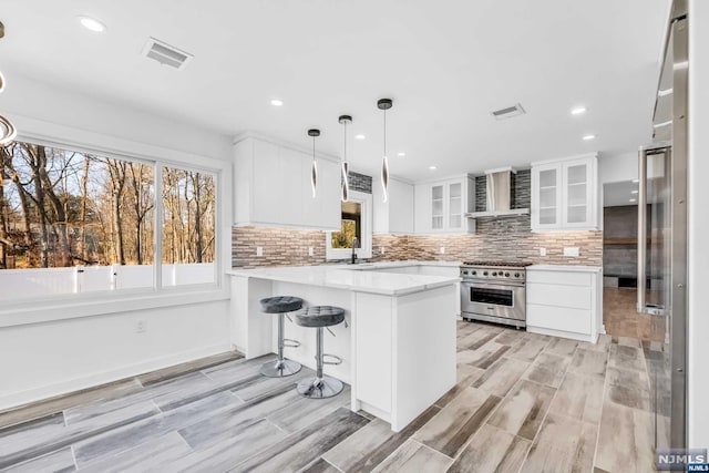 kitchen featuring kitchen peninsula, wall chimney exhaust hood, decorative light fixtures, high end stainless steel range, and white cabinetry