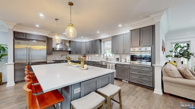 kitchen featuring a breakfast bar, hanging light fixtures, wall chimney exhaust hood, light wood-type flooring, and appliances with stainless steel finishes