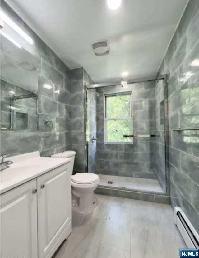 bathroom with a tile shower, a baseboard radiator, tile walls, vanity, and toilet