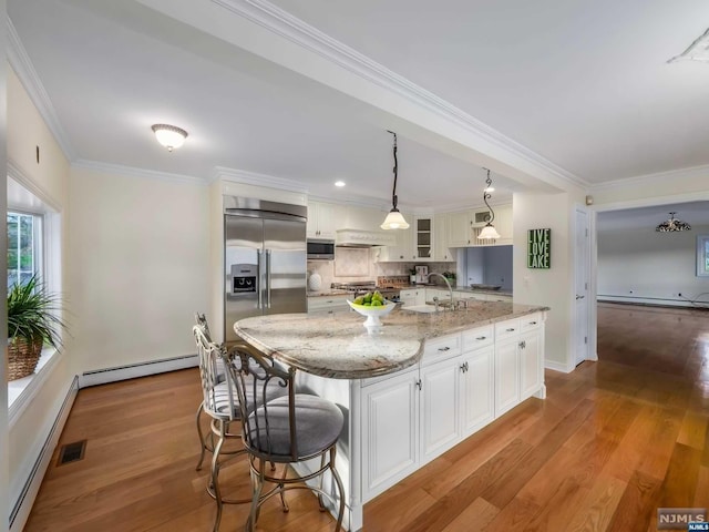 kitchen with white cabinets, hanging light fixtures, baseboard heating, appliances with stainless steel finishes, and an island with sink