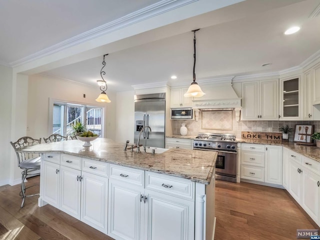 kitchen featuring dark hardwood / wood-style flooring, custom range hood, a kitchen island with sink, built in appliances, and hanging light fixtures