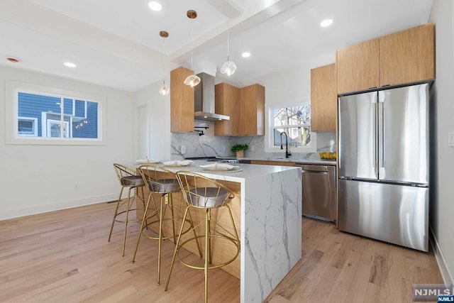 kitchen featuring hanging light fixtures, wall chimney range hood, light stone counters, light hardwood / wood-style floors, and appliances with stainless steel finishes