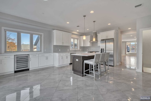 kitchen featuring appliances with stainless steel finishes, a kitchen island, wall chimney range hood, decorative light fixtures, and wine cooler