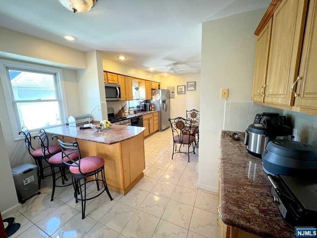 kitchen with a kitchen bar, kitchen peninsula, stainless steel appliances, ceiling fan, and dark stone countertops